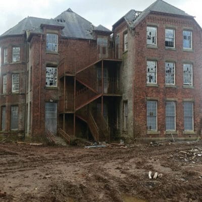 A large, derelict red-brick hospital building with broken windows and an external metal fire escape at the Whittingham Hospital site, surrounded by muddy ground and debris during a clearance project.