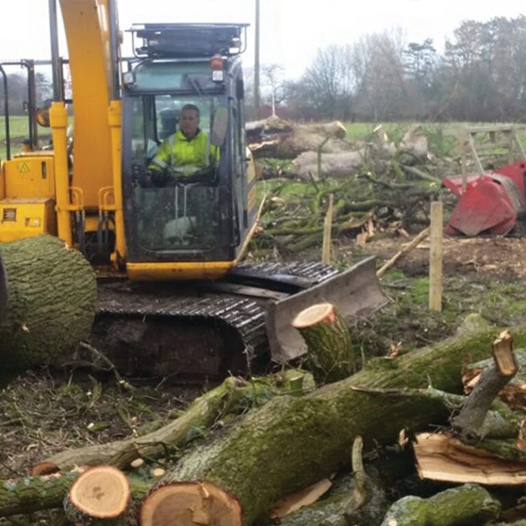 A yellow excavator being operated by a Highfield Tree Care professional to move and clear large felled tree trunks on a muddy development site.