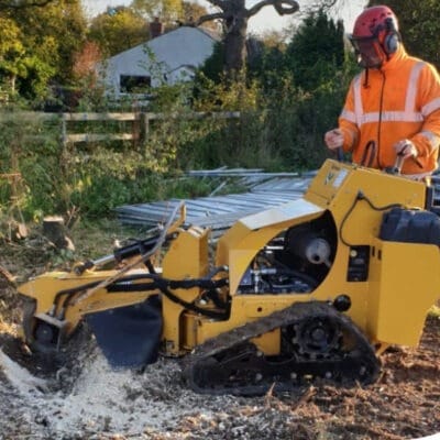 A powerful yellow Predator P4012 stump grinder being used by Highfield Tree Care to clear large tree stumps on a development site, with a company van and greenery in the background.
